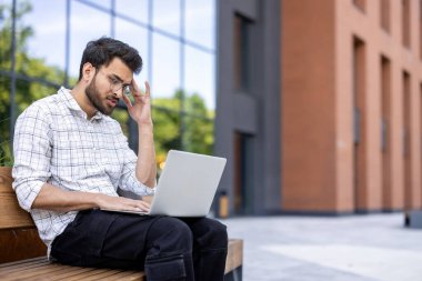 Endişeli genç Hintli bir adam dışarıda bir bankta oturuyor, dizüstü bilgisayarını kucağında tutuyor, ekranda hayal kırıklığına uğramış görünüyor ve eliyle kafasını tutuyor..