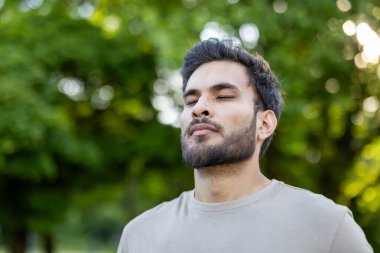 Genç adam kapalı gözlerle ayakta duruyor. Açık hava ortamında sükuneti kucaklıyor. Etrafı yemyeşil ağaçlarla çevrili, doğal ortamda sükuneti ve huzuru yansıtıyor..