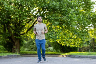 Genç adam yeşil ağaçlarla çevrili dışarıda koşuyor, müzik dinliyor. Güneşli bir günde spor ve doğanın tadını çıkarmak. Pozitif enerji ve sağlıklı yaşam tarzı kavramı.