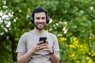 Parktaki akıllı telefona gülümseyen kulaklık takan genç bir adam. Rahat açık hava ayarı, güneşli bir günde doğa ile keyif, teknoloji, rahatlık ve bağlantı yakalar..