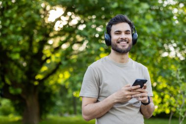 Kulaklık takan, parkta telefonu tutan gülümseyen genç bir adam. Açık havada aktifken müziğin tadını çıkarmak. Sağlıklı yaşam tarzını, teknolojiyi, keyfi temsil eder.