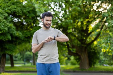 Genç adam spor yaparken dışarıda akıllı saatleri kontrol ediyor. Yeşilliklerle çevrili, park ortamında aktif yaşam tarzının tadını çıkarıyor. Teknolojiye ve doğal çevredeki egzersize odaklanın.