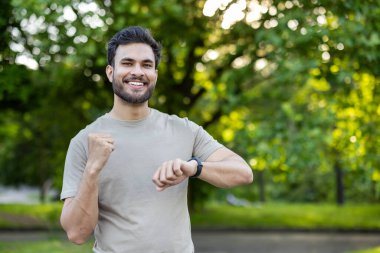 Hevesli sporcu aktif egzersiz seansından sonra bileğindeki spor takip cihazını kontrol ediyor. Gündelik kıyafet ve neşeli ifade, yemyeşil bir ortamda tatmin ve canlılık iletir..