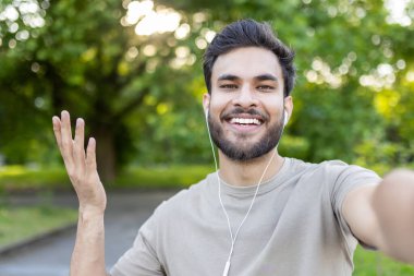 Genç adam parkta akıllı telefon ve kulaklıkla selfie çekerken gülümsüyor. Boş zamanlarında mutluluk, bağlantı ve rahatlamış açık hava atmosferini yakalar..