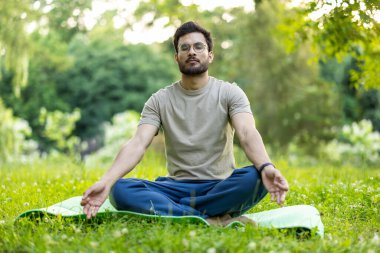 Yeşilliklerle çevrili yemyeşil parkta meditasyon yapan bir adam. Sakin ve odaklanmış ifade, yoga minderinde bağdaş kurmuş oturuyor. Farkındalık, rahatlama, huzur ve açık hava refahı kavramları için mükemmel..