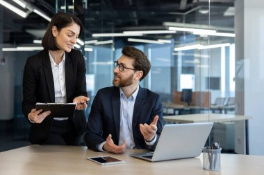 Modern ofiste iş arkadaşları dizüstü bilgisayarla tartışıyorlar. Elinde belgeler olan bir kadın, oturan bir adam. Etkileşim, takım çalışması, profesyonellik, şirket ortamı.