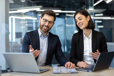 İş adamları modern ofiste dizüstü bilgisayarlar kullanarak ortak bir tartışmaya giriştiler. Fikirlerini paylaşıp proje üzerinde çalışan iki meslektaş. Gülümseme etkileşimi takım çalışmasını vurgular