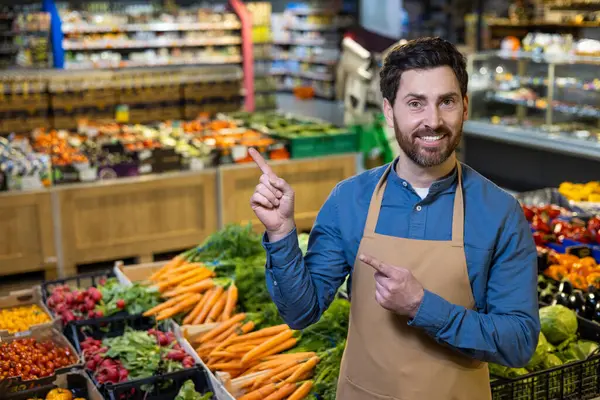 Önlüklü gülümseyen market çalışanı rengarenk taze ürünler bölümüne gururla işaret ediyor. Sağlıklı yaşam tarzını, beslenmeyi simgeleyen çeşitli meyve ve sebzelerin gösterimi