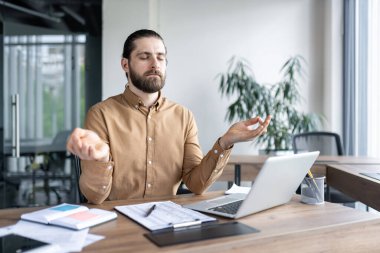 Dengeyi bulmak ve stresi azaltmak için çalışma masasında meditasyon yapan rahat profesyonel bir adam. Laptop ve belgelerle modern ofis ortamında oturan, mesai saatlerinde farkındalığı kucaklayan.