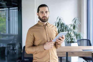 Genç adam elinde tabletle modern ofiste kendinden emin duruyor. Profesyonel iş ortamı, teknoloji ve liderlik kavramı. Odaklanma ve kararlılığı gösterir.