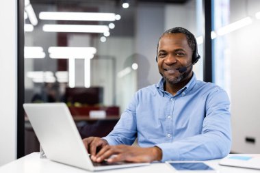 Ofis ortamında dizüstü bilgisayarla çalışan Afro-Amerikalı olgun iş adamı. Profesyonel iletişim için kulaklık takıyor. Parlak ve modern çalışma alanı profesyonelliği ve bağlılığı iletir.