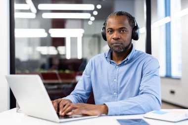 Kulaklık takan Afro-Amerikalı yetişkin iş adamı laptopu kullanarak görevlere odaklandı. Profesyonel ortam ofis çalışma alanını gösteriyor. Konsantrasyonun ifadesi