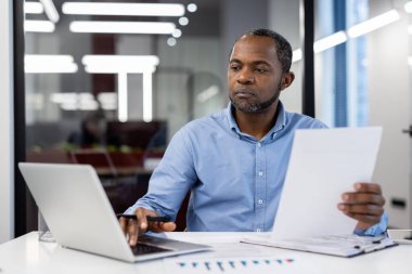 Afro-Amerikalı olgun iş adamı masasında işine odaklanıyor. Kağıtları tutmak, modern ofiste dizüstü bilgisayar kullanmak. İş ortamında odaklanma, adanmışlık ve profesyonellik göstergesi.