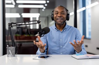 Afro-Amerikalı olgun iş adamı kulaklık ve mikrofon kullanarak modern ofis ortamında podcast yapıyor. Bağlı görünüyor, anlamlı jestlerle içeriği tartışıyor..