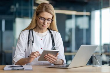 Tıp bürosunda steteskoplu, tablet ve laptop kullanan beyaz önlüklü bir doktor. Sağlık teknolojisi ve hasta veri yönetimi üzerine odaklanın.