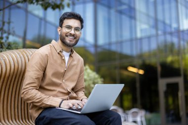 Sakallı ve gözlüklü genç adam modern binanın dışındaki bankta dizüstü bilgisayar kullanarak oturuyor. Gülümsüyor, mutluluk ve verimlilik taşıyor. Günlük giysiler, uzak mesafelere daha rahat yaklaşmamızı öneriyor..