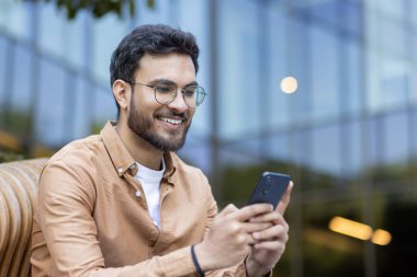 Genç adam dışarıda oturmuş mutlu bir ifadeyle telefonu kullanıyor. Akıllı telefonla uğraşırken gülümsüyor, gözlük takıyor ve günlük gömlekler giyiyor. Arka plandaki modern mimari görüntüye kentsel hissi ekler.