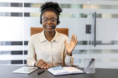 Kendine güvenen genç Afrikalı Amerikalı kadın sanal görüşme için kulaklık kullanıyor. Gülümseme ve el sallama, elinde tablet, defter, profesyonelliği ve arkadaşlığı yansıtan ofis masası..