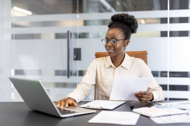 Masadaki kendine güvenen genç Afrikalı Amerikalı kadın dizüstü bilgisayar kullanıyor ve belgeleri inceliyor. Modern ofis ortamında verimlilik, odak ve profesyonel çalışma ortamı sunar.
