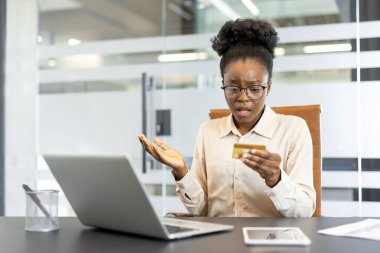 Ofisteki genç Afrikalı Amerikalı kadın kredi kartı tutarken stresli görünüyor. Bilgisayarının başında endişeli görünüyor, maddi sorunları veya ödeme sorunlarını sembolize ediyor..