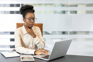Genç Afro-Amerikan kadın ofis masasında oturmuş dizüstü bilgisayarla çalışıyor boğazı ağrıyor. Odaklanma, mesleki ortamda çoklu görev yaparken endişe ve rahatsızlığı gösterir