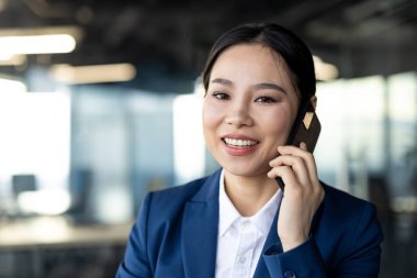 Asyalı iş kadını akıllı telefonla konuşurken gülümsüyor, modern ofiste mavi takım elbise giyiyor. Resim, profesyonel başarı, iletişim ve pozitifliği iletir. İş ve teknoloji için uygun.