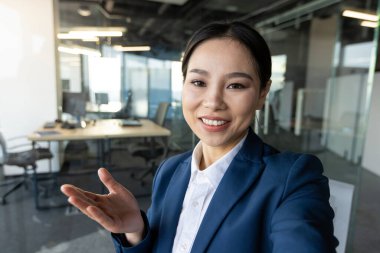 Mavi takım elbiseli kendine güvenen bir kadın, modern ofis ortamında içten bir gülümsemeyle video çağrısına katılıyor. Sanal iletişimi geliştirerek profesyonelliği ve ulaşılabilirliği hedefliyor..
