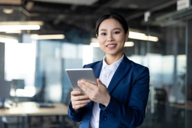 Asyalı iş kadını ofiste tablet kullanıyor. Olumlu ifadeli profesyonel kıyafetler başarıyı yansıtır. Dijital strateji, sergilenen teknoloji ve modern işyeri dinamikleri ile meşgul