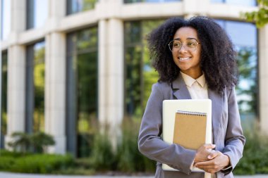 Ofis binasının önünde elinde not defteri ve tabletle duran, kendine güvenen, gözlüklü genç öğretmen. Gülümseyen, profesyonel ve doğal aydınlatma, başarı ve pozitiflik içinde rahatlamış..