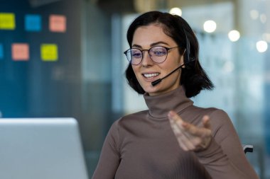 İş kadını internetteki toplantıda kulaklık takıyor. Modern ofis ortamında sanal sohbet sırasında gülümsemek. Müşteri hizmetlerine odaklı profesyonel ayar