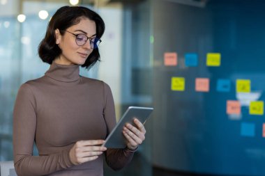 İş kadını, ofiste iş faaliyetlerinde bulunan bir tablet tutuyor. Planlama ve organizasyon için teknolojiyi kullanan, gözlük takan ve günlük giysiler giyen kendine güvenen bir profesyonel..