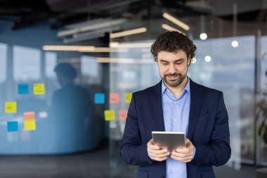 Mavi takım elbiseli bir iş adamı modern ofiste tablet kullanıyor. Gülümseyen ve odaklanmış halde, renkli yapışkan notlarla cam duvarın yanında duruyor, verimli çalışma atmosferini somutlaştırıyor..