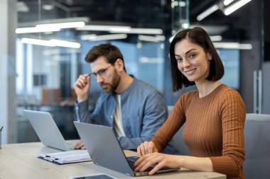 Modern ofis ortamında iş birliği yapan iki profesyonel. Dizüstü bilgisayarda yazarken gülümseyen bir kadın, dizüstü bilgisayarda düşünceli bir ifadeyle çalışan bir adam.