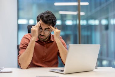 İş adamı iş yerinde stres ve yorgunluk yaşıyor, iki eliyle başını tutuyor. Ofis ayarları arasında, profesyonel çevredeki baskı ve hayal kırıklığını yansıtan dizüstü bilgisayar yer alıyor.