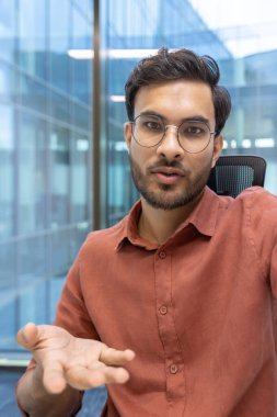 Gözlüklü bir iş adamı modern ofiste el kol hareketi yaparak video çağrısında bulunuyor. Profesyonel iletişim ve telefon aracılığıyla iletişim. Kendinden emin ifade, odaklı konuşma