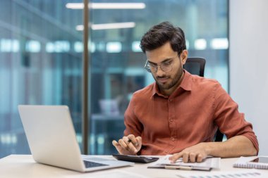 Ofiste hesap makinesi ve dizüstü bilgisayar kullanarak çalışan genç bir girişimci. Ciddi ve finansal hesaplamalara odaklanmış, modern işyerindeki kapalı mekanlarda oturuyor. İş ve verimliliği temsil eder