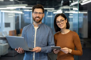 Modern ofiste dizüstü bilgisayar ve tabletle birlikte poz veren iş adamları. Erkek ve kadın gülümsüyor, takım çalışmasını, özgüveni ve başarılı işbirliğini temsil ediyor..