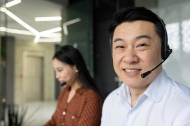 Ofisteki Asyalı iş adamları iletişim için kulaklık kullanıyor. Müşteri hizmetlerinde çalışırken kameraya gülümseyen bir adam. Takım çalışmasını ve desteği vurgular