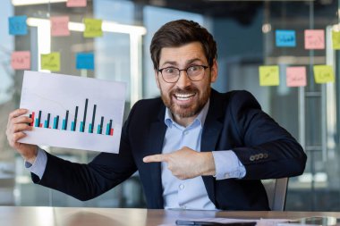 Kendine güvenen işadamı, iş toplantısı sırasında satış grafiğini coşkuyla sunuyor. Mesleki kıyafet, olumlu ifade, ofis ortamında başarı ve başarının altını çiziyor.