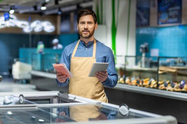 Önlük giyen tezgahtar süpermarkette elinde tablet ve paketlenmiş et tutuyor. Resim, envanter yönetimi, perakende teknolojisi ve modern market ortamını ön plana çıkarıyor