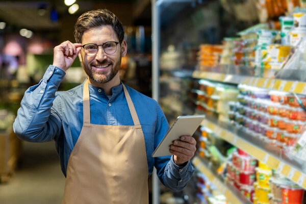 Male store employee smiling confidently while holding tablet in grocery aisle. Worker wearing apron and glasses in supermarket setting, showcasing customer service and technology use.