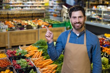 Gülümseyen market çalışanları market ortamındaki taze sebzelere işaret ediyor. Sergide havuç, yeşillik ve diğer ürünler yer alıyor. Ortamı hoş karşılamak sağlıklı seçenekleri ve müşteri hizmetlerini vurguluyor.