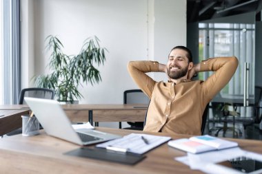İşadamı ellerini başının arkasına koyup ofiste dinleniyor. Modern çalışma alanı dizüstü bilgisayar, dokümanlar, kapalı alandaki bitkiler. Sahne, iş yeri rahatlığını, stres rahatlığını, mesleki tatmini taşır.