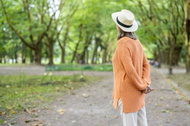 Olgun kadın yeşil ağaçlarla çevrili parkta rahatça yürüyor. Doğada sükuneti kucaklayan bir şapka ve kazak giyiyor. Sakin bir yaşam tarzı ve emeklilik neşesinin mükemmel bir tasviri..