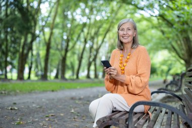 Yeşillik park ortamında akıllı telefonunu kullanarak sırıtan olgun bir kadın. Açık hava eğlencesi ve modern bağlantının özünü yakalar. Huzurlu ortamda doğa ve teknolojinin harmanlanmasını sağlar.