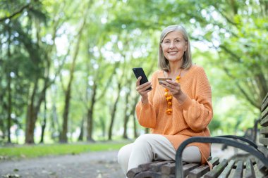 Parktaki bankta oturan gülümseyen yaşlı kadın akıllı telefon kullanıyor, kredi kartı tutuyor, dışarıda eğleniyor. Yaşlılar arasında modern yaşam tarzı ve teknoloji kullanımını gösteriyor