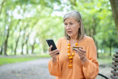 Yaşlı kadın dışarıda akıllı telefon kullanıyor, kredi kartı tutarken şok geçiriyor. Güneşli park ortamında sürprizler sergiliyor. Finansal ya da beklenmedik haberler veriyor..