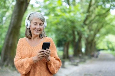 Turuncu kazaklı, kulaklık takan, ağaçlık yolda akıllı telefon kullanan yaşlı bir kadın. Olgun yaş için rahat yaşam tarzı, teknoloji ve bağlantıyı temsil eder..