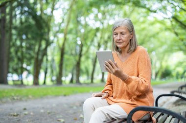 Yaşlı kadın park bankında oturmuş tablet kullanıyor, dijital teknolojiyle uğraşıyor. Dış mekan, modern teknolojiyi doğayla harmanlayarak huzurlu bir yaşam tarzını ön plana çıkarıyor..