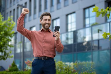 Akıllı telefona bakan neşeli iş adamı şehir sokaklarında başarıyı kutluyor. Olgun profesyonellerin modern ofis binasının dışındaki heyecanı ifade etmesi, başarıyı ve başarıyı temsil ediyor.